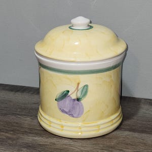 May include: A ceramic kitchen canister with a lid. The canister is painted yellow with a watercolor effect and features a green and white band around the middle. It is decorated with purple plums and green leaves.