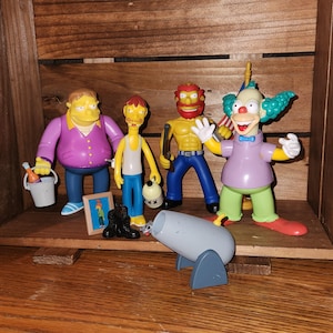 May include: A collection of Simpsons character figurines, including Barney Gumble, Sideshow Bob, Krusty the Clown, and others. The figures are displayed on a wooden shelf, with a toy cannon in the foreground. The characters are in various poses and outfits.