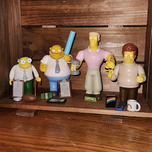 May include: Four collectible figurines of characters from The Simpsons. The figures are in various poses and outfits, with accessories like a clipboard, money, and a dumbbell. The figures are displayed inside a wooden crate.