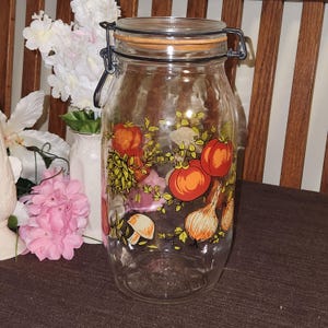May include: A clear glass jar with a hinged lid and a decorative illustration of tomatoes, onions, and mushrooms. The jar has a metal clasp and a wooden-toned lid. The illustration features red, orange, and yellow colors.