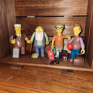 May include: Four yellow-skinned action figures from a cartoon series are displayed inside a wooden crate. The figures include a man with a microphone, an elderly man with a cane, a man with glasses, and a man holding a ball. Accessories include a phone and a briefcase.