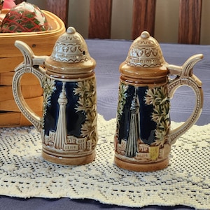 May include: A pair of ceramic salt and pepper shakers shaped like miniature beer steins. The steins are tan and brown with a blue background featuring a cityscape and a tall tower. Each stein has a handle and a decorative lid.