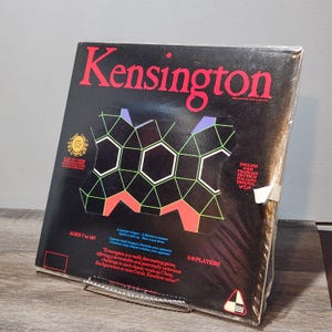 May include: A boxed board game titled "Kensington" with a black background and geometric shapes in white, green, and red. The game is for 2-6 players, ages 7 to 107. The box includes text in English, French, German, Italian, Spanish, and Arabic.