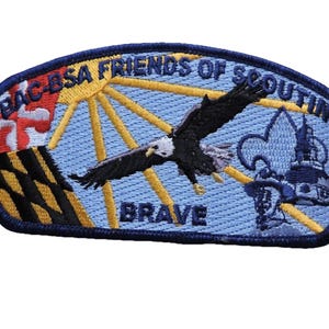 May include: Embroidered patch with a blue background featuring a soaring eagle, a sunburst, and a fleur-de-lis. Text includes "BAC BSA FRIENDS OF SCOUTING" and "BRAVE". The patch has a navy blue border.