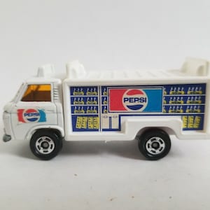 May include: White toy truck with a Pepsi logo and a side panel filled with images of Pepsi cans. The truck has a yellow tinted window and black wheels.
