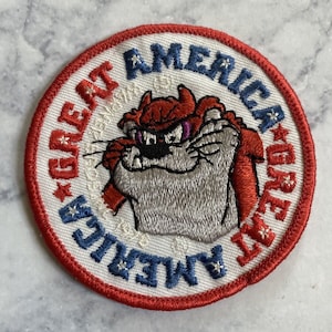 Pode incluir: Um remendo bordado vermelho, branco e azul com um desenho animado do Diabo da Tasmânia. O remendo diz "Great America" e "Great America" em um círculo ao redor do Diabo da Tasmânia.