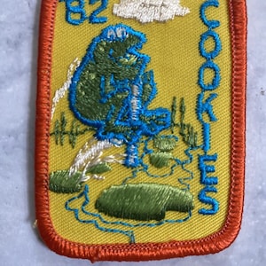 May include: A yellow embroidered patch with a blue and green cartoon character holding a cookie. The patch has the text "82 COOKIES" on it.
