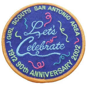Puede incluir: Parche redondo azul con ribete dorado, bordado con las palabras «Let's Celebrate» y «Girl Scouts San Antonio Area 2002». El parche conmemora el 90 aniversario, con espirales de colores.