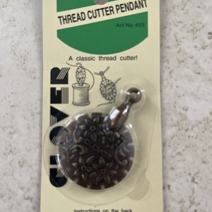 Peut inclure: Un pendentif coupe-fil doré avec un motif floral. L'emballage indique "Clover Thread Cutter Pendant Art No. 455" et "Un coupe-fil classique !"