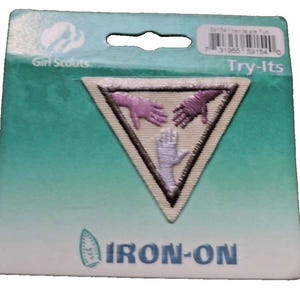 Puede incluir: Parche termoadhesivo Girl Scouts Try-Its. El parche triangular presenta tres manos bordadas en morado y blanco, que simbolizan la unidad. Se encuentra sobre un fondo degradado turquesa y blanco.