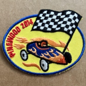 May include: Embroidered patch with a yellow oval background, featuring a blue and orange race car with a checkered flag. The text "PINewood 2014" is in red around the oval. The patch has a blue border and is likely for a racing event.