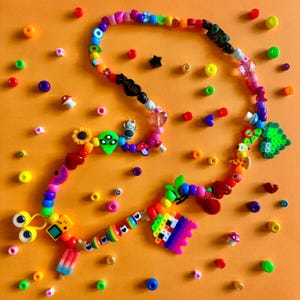 May include: A colorful beaded necklace with various charms, including a sunflower, alien, mushroom, and a popsicle. The necklace features a mix of vibrant beads in multiple colors, with a bright orange background.