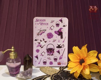 May include: A lilac sticker sheet featuring witch-themed designs such as a cauldron, witch's hat, and broom. The text "Season of the Witch" is at the top. A perfume bottle, crystal, and flowers are in the foreground.