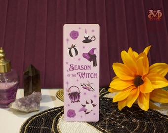 May include: A lavender bookmark with the text "Season of the Witch" and witch-themed illustrations. The design includes a pumpkin, witch hat, cauldron, and rose. A yellow flower and other decorative items are in the background.