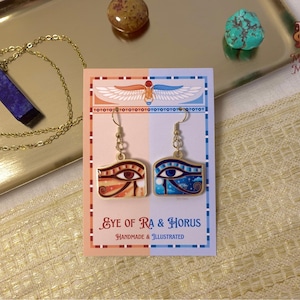 Puede incluir: Pendientes dorados con diseños del Ojo de Ra y Horus, presentados en una tarjeta decorativa. Los pendientes son de color naranja y azul, con el texto "Eye of Ra & Horus" y "Handmade & Illustrated". También se ve un collar de piedra azul.