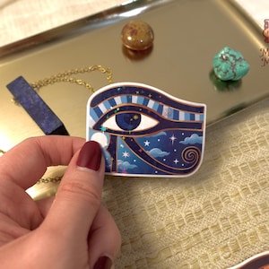 May include: A sticker featuring the Eye of Horus design, with a blue and white colour scheme, stars, and clouds. The sticker is held near a gold chain necklace and a blue crystal pendant. The background includes a gold tray and decorative stones.