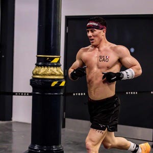 Puede incluir: Un hombre corriendo en un gimnasio, con pantalones cortos negros, guantes y una diadema con la palabra "BREAKABLE". El hombre tiene un tatuaje en el pecho y la pierna. El fondo incluye un poste negro y la palabra "MYROX".