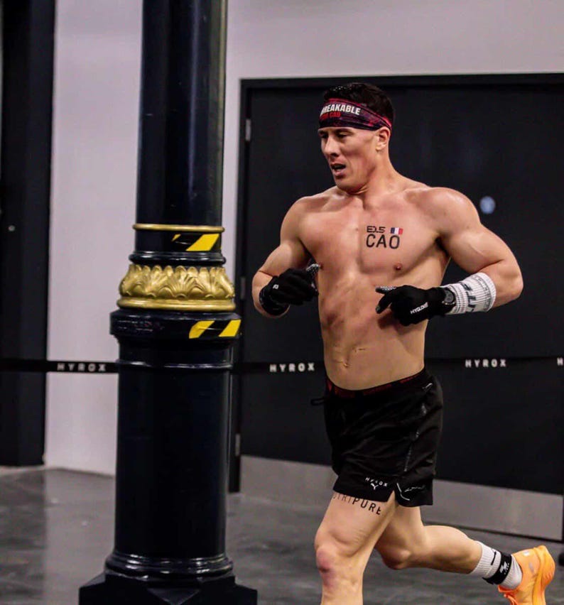 Puede incluir: Un hombre corriendo en un gimnasio, con pantalones cortos negros, guantes y una diadema con la palabra "BREAKABLE". El hombre tiene un tatuaje en el pecho y la pierna. El fondo incluye un poste negro y la palabra "MYROX".