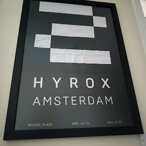 May include: A framed poster with a black border. The poster features a dark grey background with a white geometric design. The text "HYROX AMSTERDAM" is displayed in white, along with "SECOND_PLACE" and "MEN_16-24".