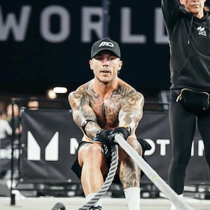 Puede incluir: Una persona con gorra y pantalones cortos negros tira de una cuerda gruesa durante un entrenamiento. La persona est&aacute; cubierta de tatuajes y lleva guantes y zapatillas deportivas. Una segunda persona est&aacute; en el fondo, con una sudadera con capucha y leggings negros.