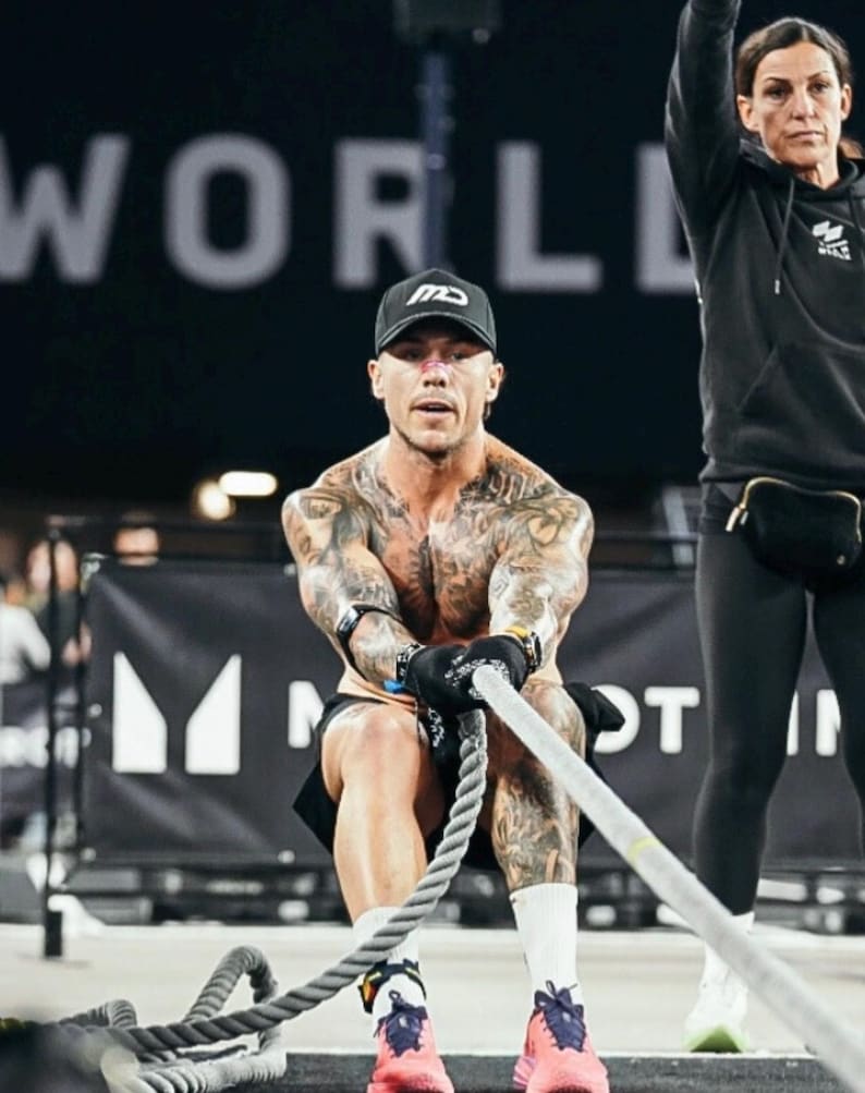Puede incluir: Una persona con gorra y pantalones cortos negros tira de una cuerda gruesa durante un entrenamiento. La persona est&aacute; cubierta de tatuajes y lleva guantes y zapatillas deportivas. Una segunda persona est&aacute; en el fondo, con una sudadera con capucha y leggings negros.