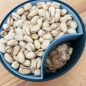 Może przedstawiać: Podzielona miska wypełniona pistacjami. Miska jest szara i ma czarną krawędź. Pistacje są w łupinach i bez łupin.