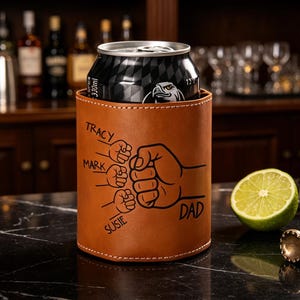 May include: A brown leather drinks can cooler with the word "DAD" and the names "Tracy", "Mark", and "Susie" printed on it. A black can sits inside the cooler. Sunglasses, a lime, and a bottle opener are also on the counter.