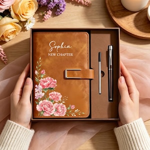 Op de afbeelding: Een lichtbruin notitieboek in lederlook met een goudkleurige metalen sluiting, een bloemenprint van roze anjers en de tekst "Sophia NEW CHAPTER" in het wit gedrukt. Het notitieboek zit in een doos met een bijpassende metalen pen en vulling.