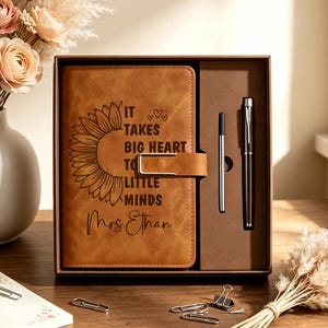 Puede incluir: Un diario de piel sintética color canela con un gráfico de girasol y el texto "IT TAKES BIG HEART TO SHAPE LITTLE MINDS" y "Mrs. Ethan" grabado en la portada. El diario está dentro de una caja de regalo marrón con un bolígrafo y un lápiz óptico.