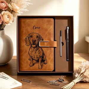 May include: A tan leather-effect journal with a metal clasp, featuring an engraved illustration of a dog and the name Coco. The journal is presented in a brown gift box alongside a matching pen and a thin metal stylus, all set in a custom tray.