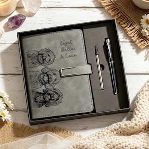 May include: A grey notebook with a magnetic strap closure, featuring an engraved illustration of three dogs and the text Eigel, Bella & Coco. The notebook is presented in a gift box alongside a black pen and a silver metal pen refill cartridge.