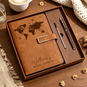 Könnte beinhalten: Ein reisejournal aus hellbraunem kunstleder in einer braunen box, mit einer weltkarten-gravur, einem goldenen metallverschluss und dem text "William's TRAVEL JOURNAL". Das set enthält einen dunkelbraunen stift und eine silberne mine.