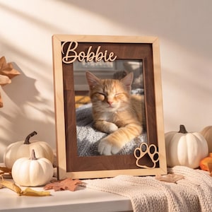 May include: A rectangular wooden picture frame featuring a light-colored outer border and a dark-colored inner frame. The name Bobbie is cut out in script lettering at the top, and a paw print cutout is positioned at the bottom right corner.