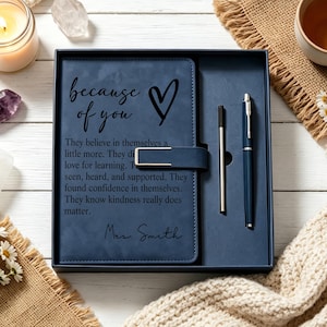 May include: A navy blue notebook set in a matching box, featuring a gold-toned magnetic clasp. The cover is engraved with a heart and the text: because of you, They believe in themselves a little more, They discover love for learning, They feel seen, heard, and supported, They found confidence in themselves, They know kindness really does matter, Mrs. Smith. A slim pen is included.