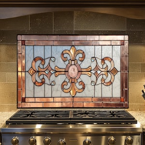 May include: A decorative stained glass panel with a copper frame and a fleur-de-lis design above a stainless steel gas range. The glass features a mix of colors, including copper, gold, and pink, with a marble-like background.