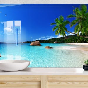 Puede incluir: Una vibrante escena de playa se muestra en un panel de vidrio, mostrando un paraíso tropical. La imagen presenta un cielo azul claro, agua turquesa, arena blanca, palmeras y una isla rocosa. Un lavabo blanco y un grifo cromado están en primer plano.