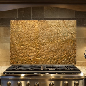May include: A kitchen backsplash with a textured, golden-brown finish. The backsplash is installed above a stainless steel gas range with black grates. The kitchen features dark wood cabinetry and a granite countertop.
