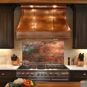 May include: A copper range hood with a hammered texture is the focal point of this kitchen. The hood is above a stainless steel gas range and a backsplash with a mottled, rust-colored finish. Dark wood cabinets flank the range.