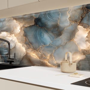 Könnte beinhalten: Eine Küchenrückwand mit einem blauen, goldenen und weißen Marmormuster. Ein schwarzer Wasserhahn und ein Waschbecken befinden sich im Vordergrund, zusammen mit einem beigen Korb und einem kleinen weißen Quadrat mit einem goldenen Kreis. Die Arbeitsplatte ist weiß.