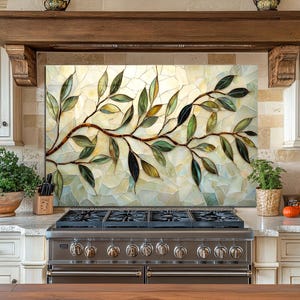 May include: A kitchen backsplash with a stained glass design featuring a brown branch with green and gold leaves. The backsplash is above a stainless steel gas range. The kitchen has light-coloured cabinets and countertops.