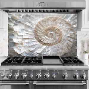May include: A kitchen splashback with a large, swirling nautilus shell design. The shell is made of iridescent, pearlescent tiles in white and tan. It's above a stainless steel gas range, a stylish kitchen feature.