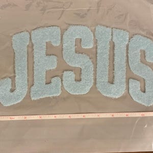 May include: Light blue, fuzzy fabric letters spell out the word "JESUS". The letters are approximately 10 inches wide. The letters are on a clear, mesh-like material, with a measuring tape in the image.