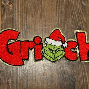 May include: A red and green Grinch patch with gold trim. The word "Grinch" is spelled out in red, textured letters. The "i" is replaced with the Grinch's face wearing a Santa hat.
