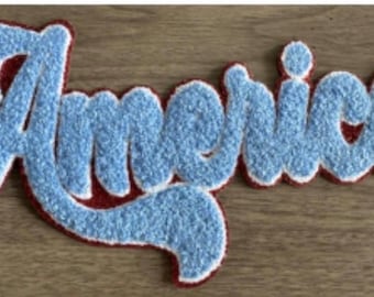 May include: A light blue, textured patch with the word "Americas" in a cursive font. The patch has a red border with a white outline, and is set against a wooden background. The patch is likely for clothing or accessories.