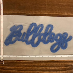May include: A blue felt applique with the word "Bulldogs" in a cursive font. The applique is approximately 11 inches long and is on a light gray mesh backing.