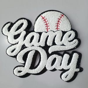 May include: A baseball-themed decoration with the words "Game Day" in white, textured lettering outlined in black. A white baseball with red stitching sits above the text. The design has a glittery finish.