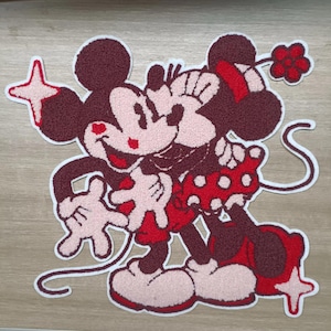 May include: A burgundy and red embroidered patch featuring Mickey and Minnie Mouse embracing. Minnie wears a red polka dot skirt and bow. Mickey holds a star. The patch has a white border.