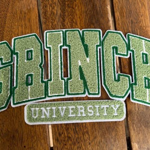 May include: Green felt patches with the word "GRINCH" in large, block letters and "UNIVERSITY" in smaller letters. The patches have a white border and are set against a wooden background.
