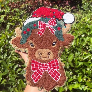 May include: A brown, textured cow-shaped Christmas ornament. The ornament features a red Santa hat, a green wreath with red berries, and two red plaid bows. The cow has a light brown face and is decorated with black eyes.