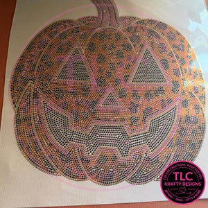 May include: A pumpkin-shaped iron-on transfer design covered in small, shimmering rhinestones. The pattern features a brown and black leopard print motif with triangular eyes and a jagged mouth. A circular logo in the corner reads TLC KRAFTY DESIGNS.